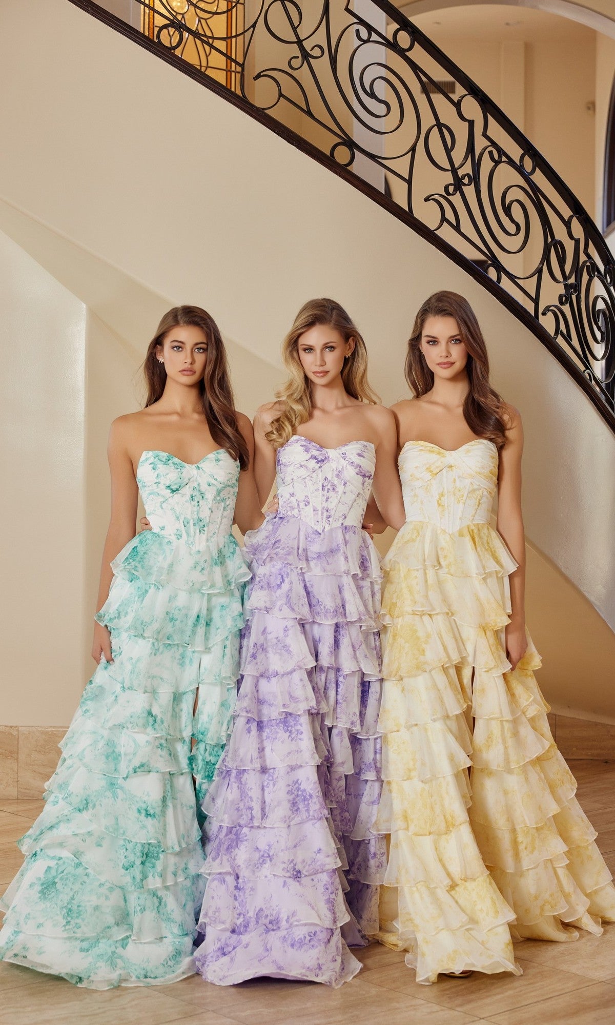 Three women stand side by side by a staircase, each wearing the Nox Anabel T1865 long prom dress—strapless, floral chiffon with an elegant a-line ruffle skirt—in pastel green, purple, and yellow patterns.
